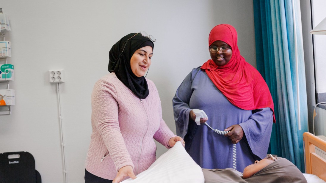 Två kvinnor vid en säng med en liggande kvinna i.