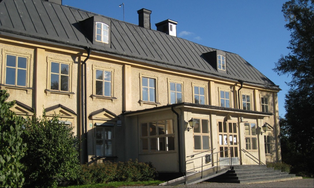 En gul herrgårdsbyggnad i solsken med blå himmel bakom.